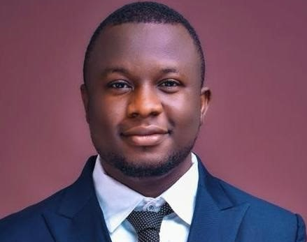 A person is dressed in a dark blue suit, white shirt, and patterned black tie against a plain pink background.