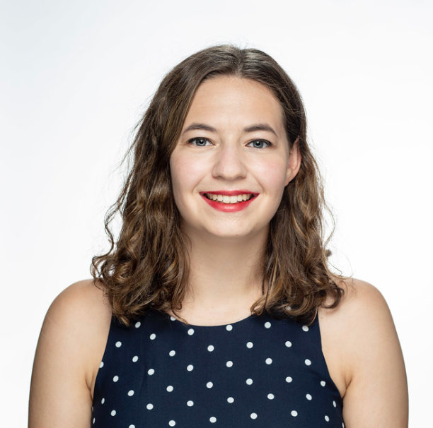 Person with shoulder-length wavy brown hair wearing a sleeveless navy top with white polka dots against a light background.