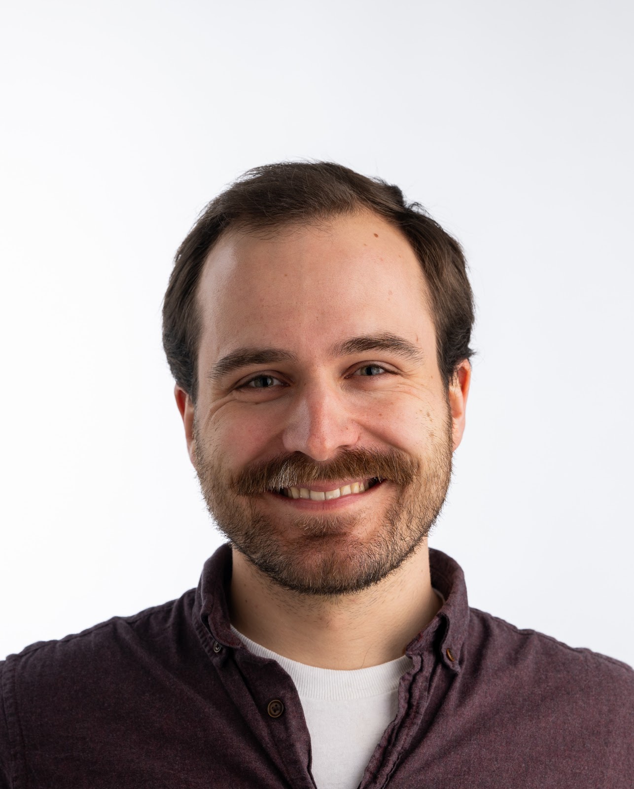 Person with brown hair wearing a maroon button-up shirt over a white crewneck t-shirt against a plain white background.